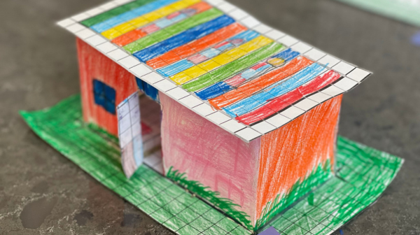 A small house made out of paper, colored and sitting on a piece of colored green paper.