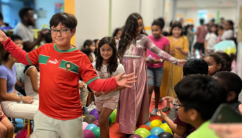 An photo of a group of kids walking down the hallway single-file, with other students sitting on either side of the hallway.