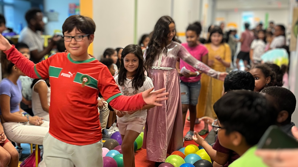 An photo of a group of kids walking down the hallway single-file, with other students sitting on either side of the hallway.