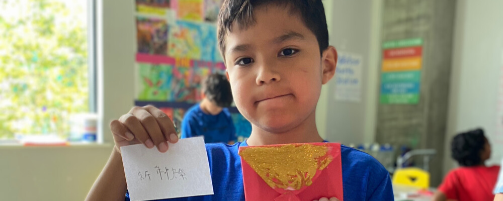 A boy holding up a red envelope with gold glitter and a white piece of paper with Chinese characters written on it.