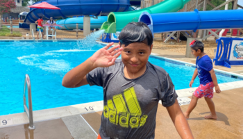 A smiling boy with wet hair and a wet shirt waving at the camera, standing in front of a pool