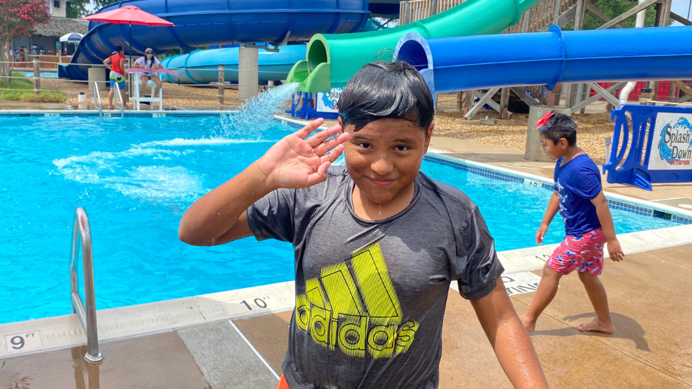 2023 Camp highlights blog featured images (1000 × 563 px)(4) A smiling boy with wet hair and a wet shirt waving at the camera, standing in front of a pool