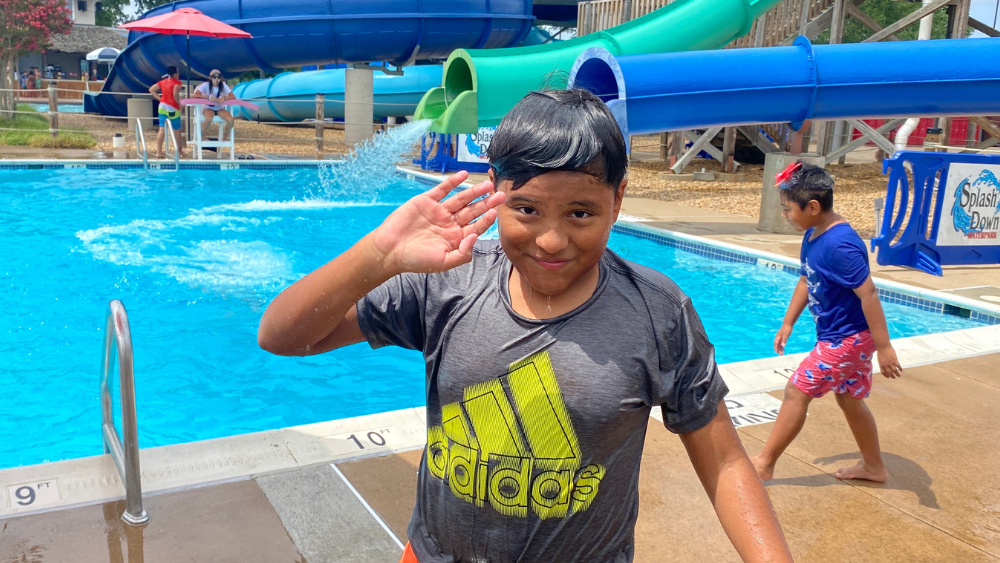 A smiling boy with wet hair and a wet shirt waving at the camera, standing in front of a pool