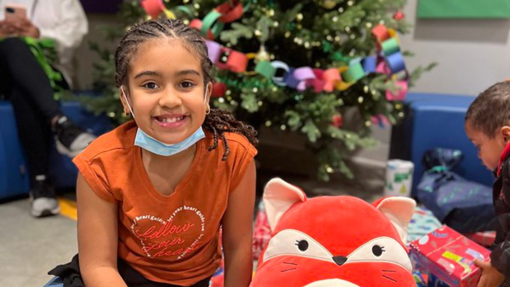FY24 Blog Post Featured Images(1) A girl smiling at the camera, sitting in front of a Christmas tree with a red stuffed animal next to her.