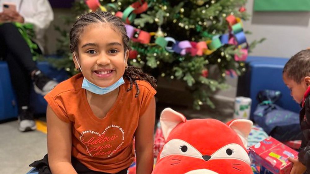 A girl smiling at the camera, sitting in front of a Christmas tree with a red stuffed animal next to her.