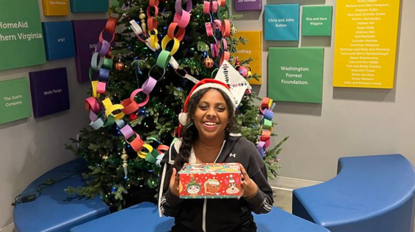 FY24 Website Event Featured Images(3) A girl holding a present, wearing a Santa hit, sitting in front of a Christmas tree and smiling