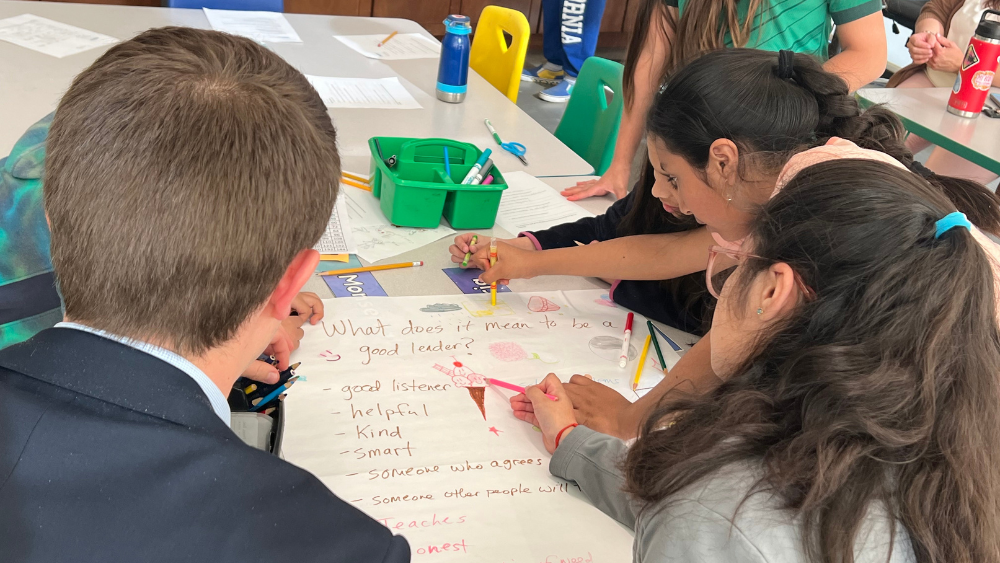 A group of students and an adult writing on a large sheet of white paper