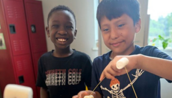 A photo of two boys smiling at the camera and constructing a tower out of spaghetti noodles and marshmallows